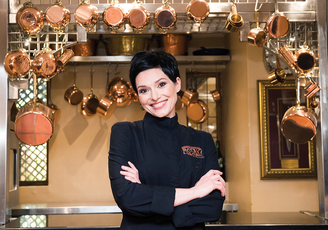 Head chef in a black tunic standing under a hanging rack of copper pans