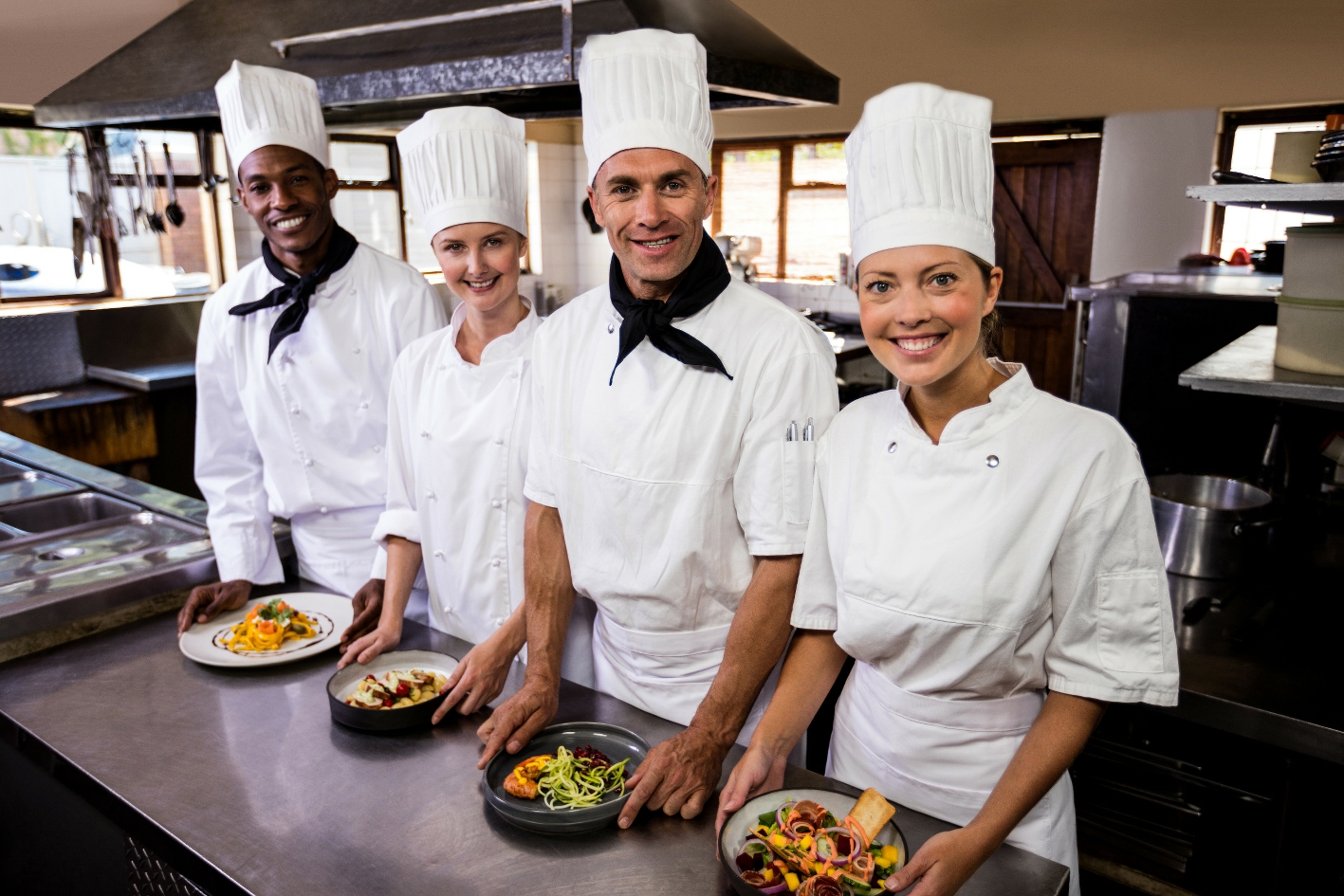 Four chefs in whites lined up at a stainless-steel pass with plated dishes in a Franschhoek kitchen