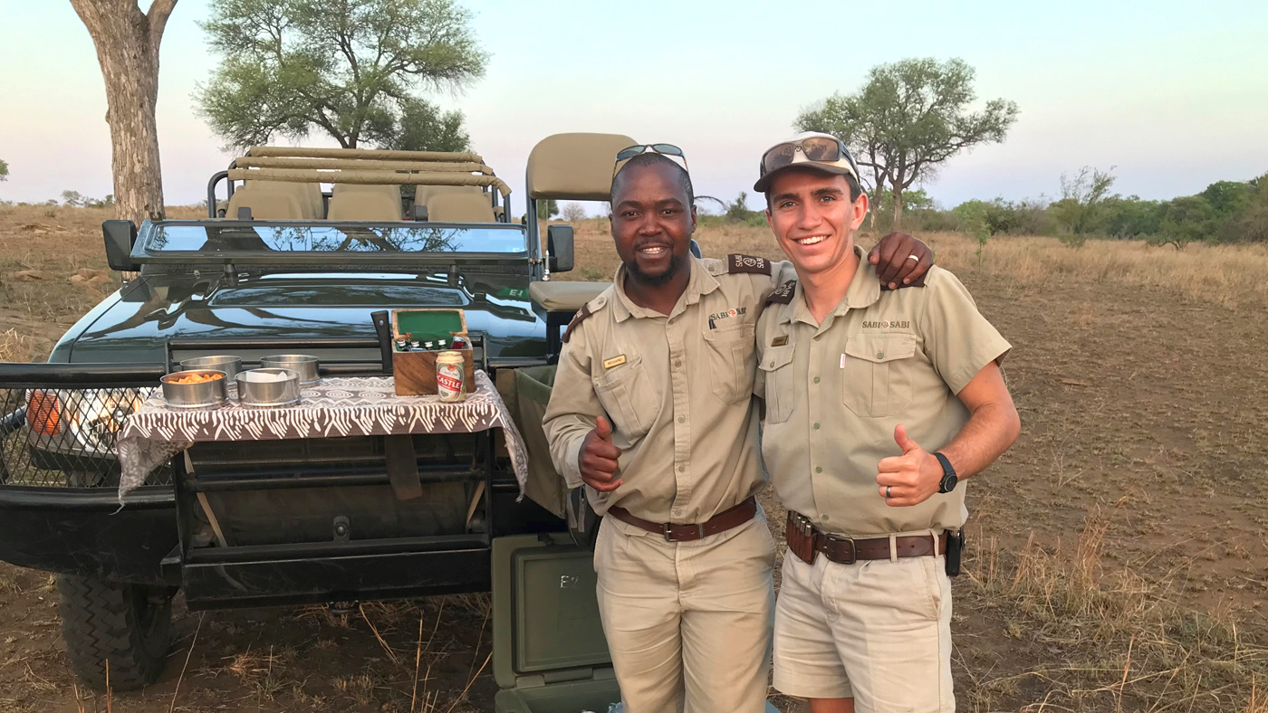 Two Sabi Sabi rangers in uniform standing in front of a game-drive vehicle giving thumbs up at sunset