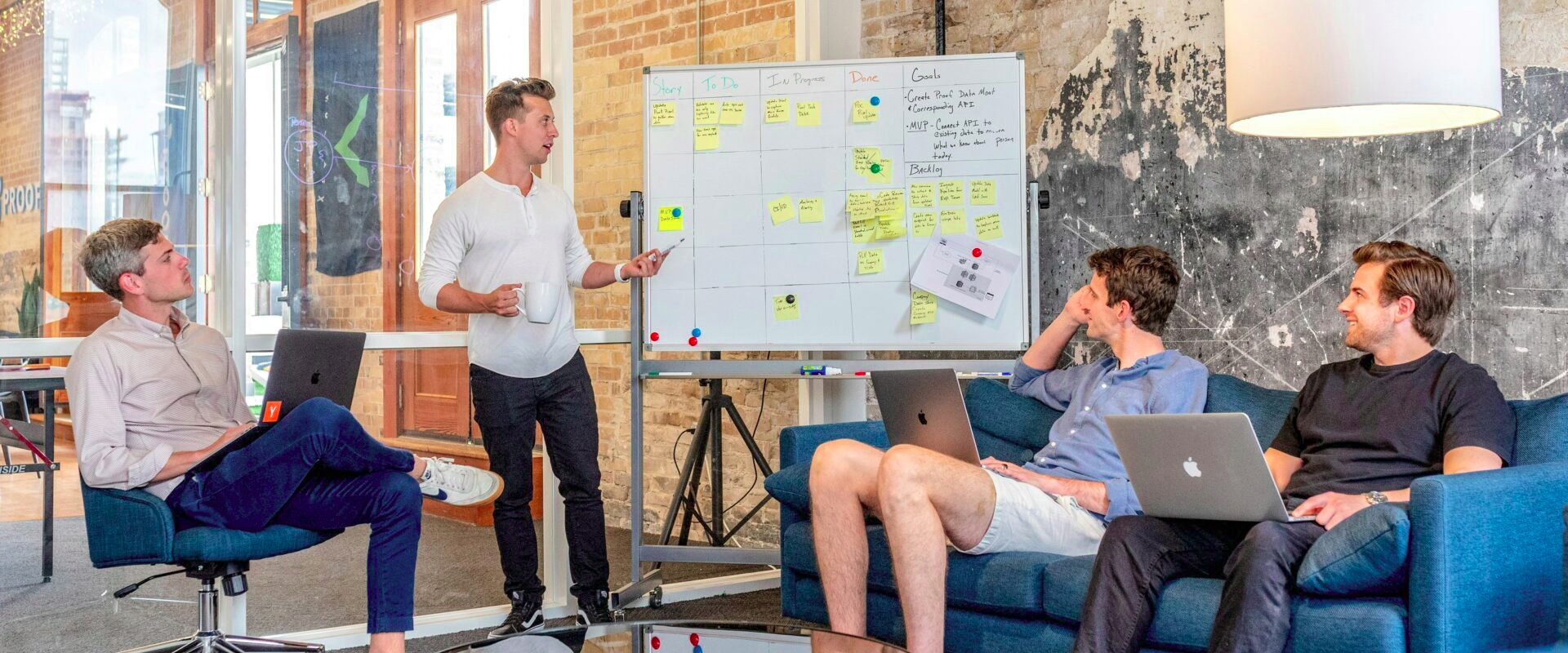 A standup at the campus whiteboard — sticky notes, scrum board, mixed cohort